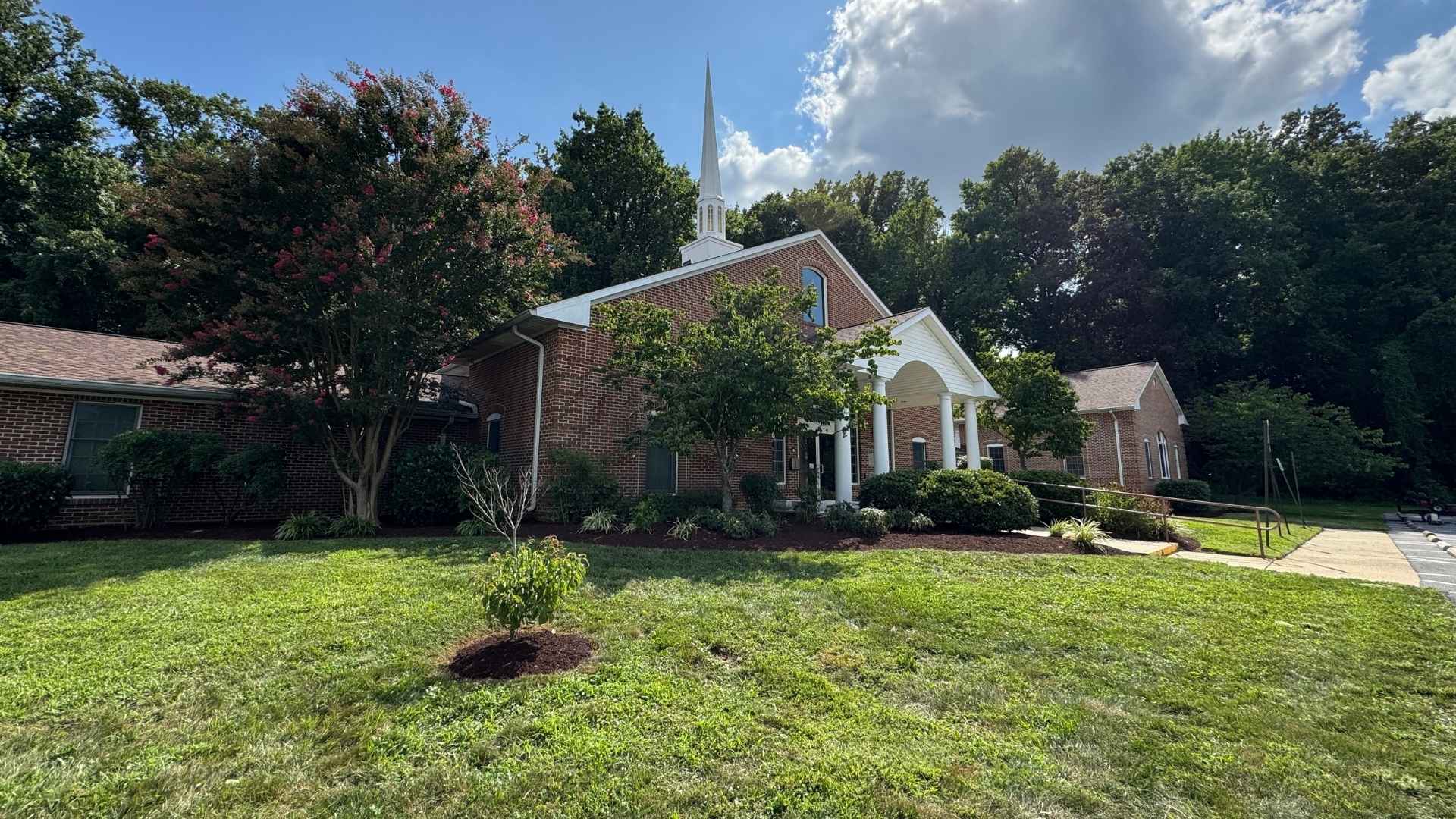 Small brick church with a steeple surrounded by trees and greenery on a sunny day.